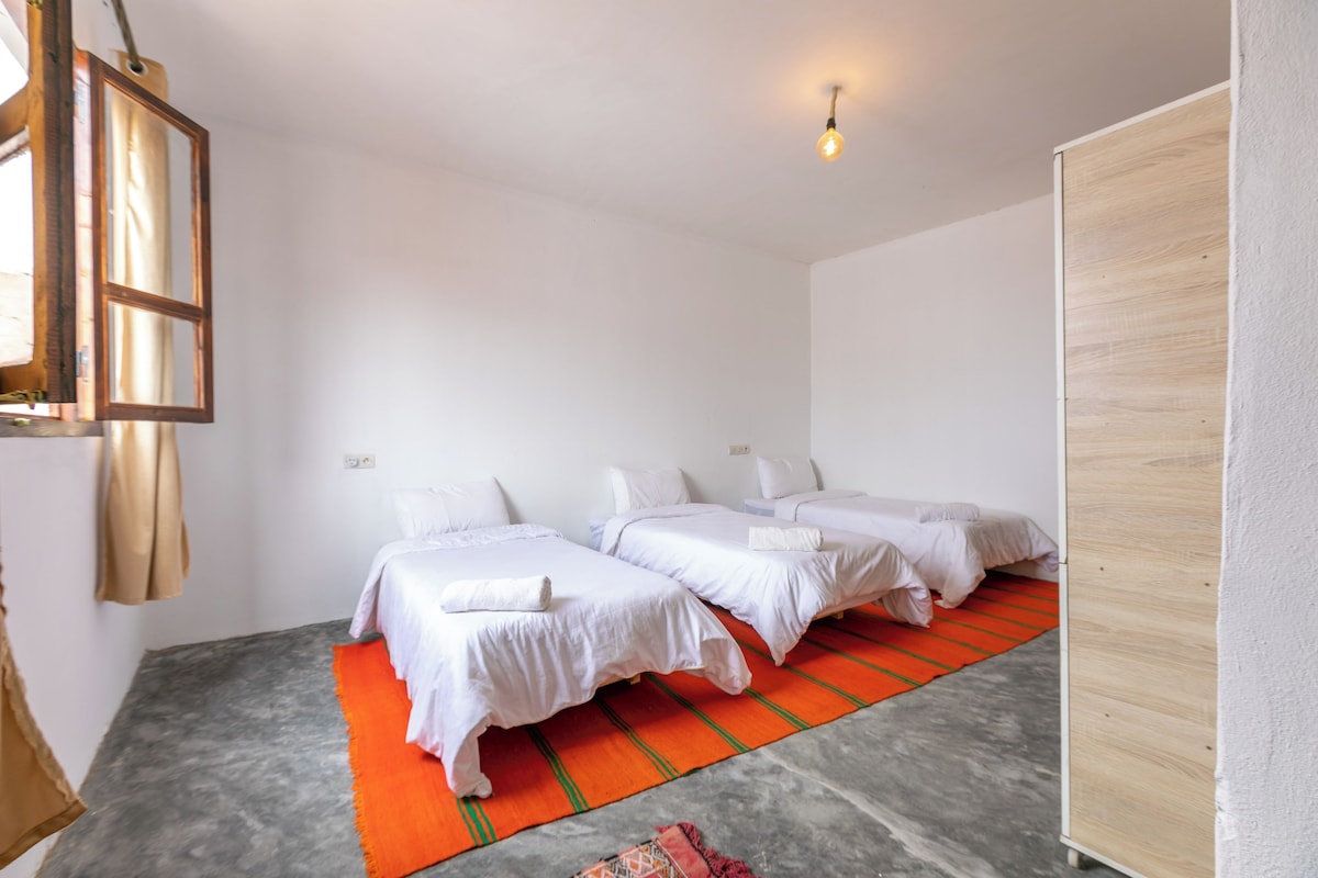 Ein Hotelzimmer mit zwei Betten, einem rot-grün gestreiften Teppich, einem hölzernen Kleiderschrank und einer Pendelleuchte.