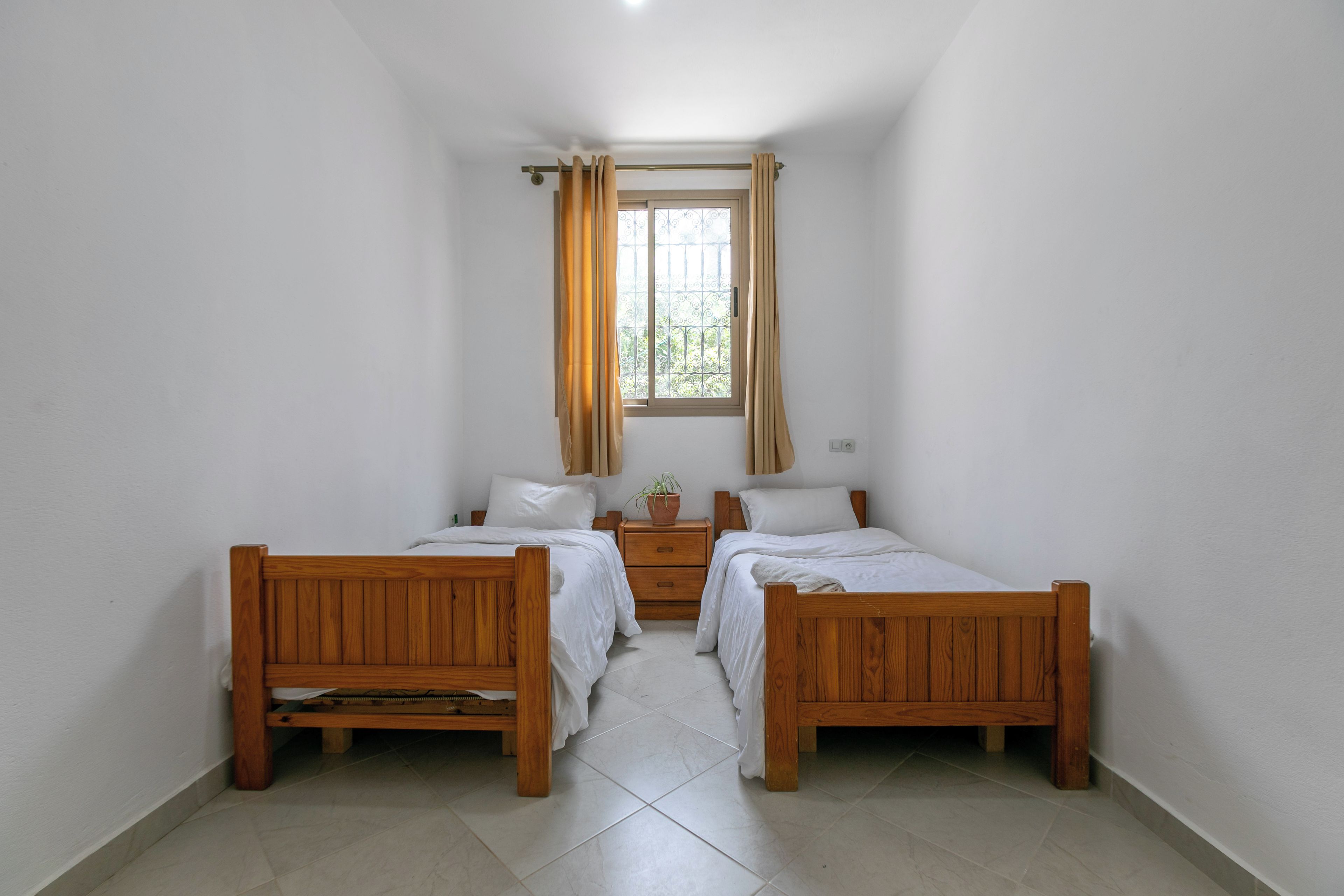A room with two wooden beds, a window with curtains, a bedside table with a plant, and a wall socket.
