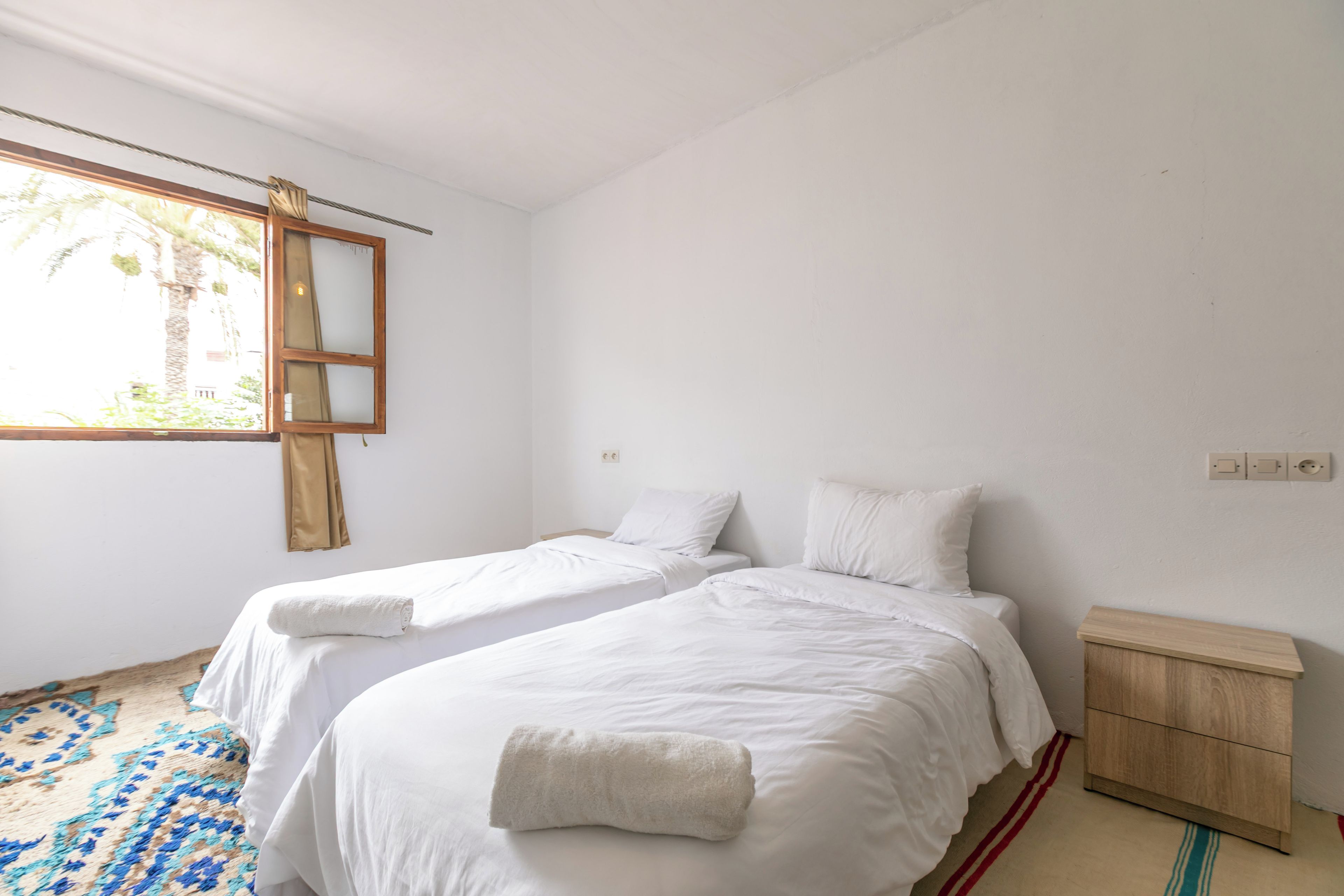 Un dormitorio con dos camas individuales, una mesita de noche de madera, un pequeño armario de madera y una ventana con cortinas.