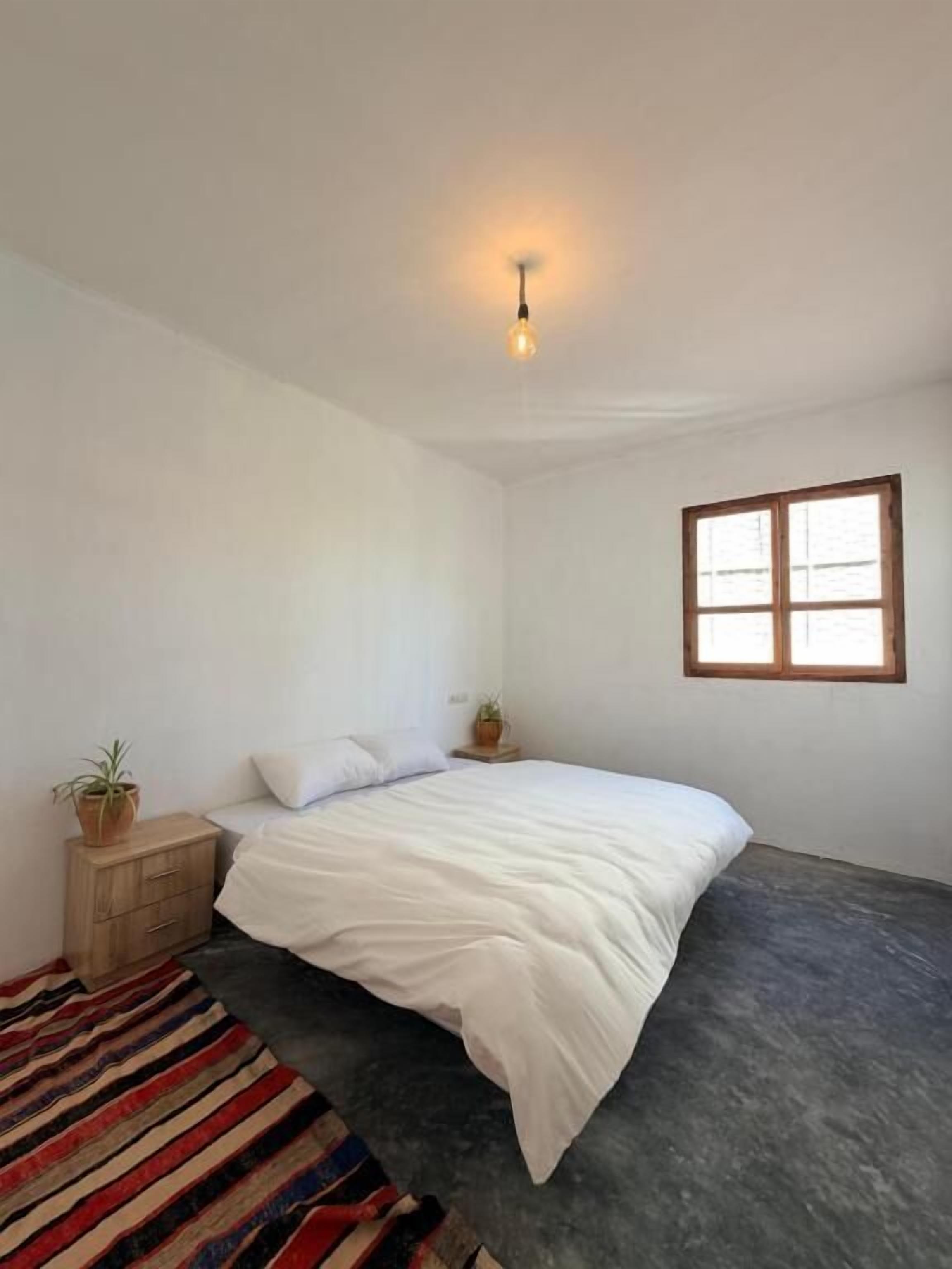 Un dormitorio con una cama, un ventanal, una lámpara y un tapete a rayas.