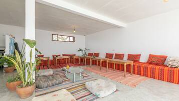 A spacious living area with a high ceiling, a long wooden sofa set, a glass coffee table, and a variety of colorful rugs.