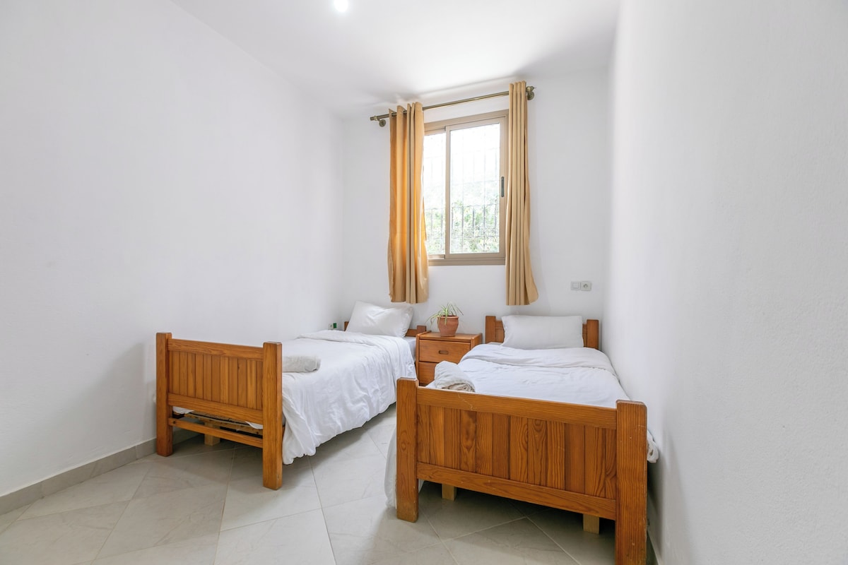 Ein Schlafzimmer mit zwei hölzernen Betten, weißer Bettwäsche, einem Fenster mit Vorhängen und einem kleinen Nachttisch mit einer Pflanze.