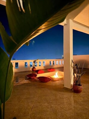 A rooftop terrace with a fire feature, potted plants, and a view of the city at dusk.