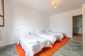 Quarto de hotel com três camas de solteiro, banheiro com pia e uma pequena janela.