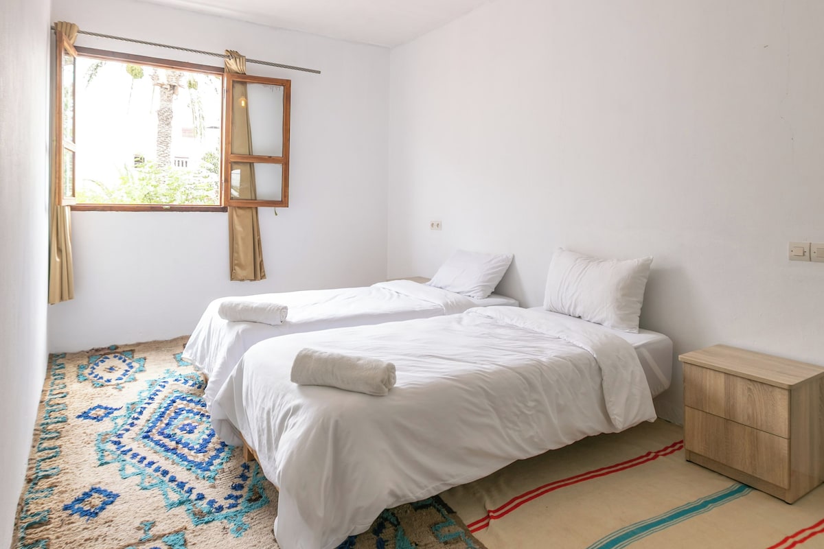 Ein Zimmer mit zwei Betten, einem fensterbelaubten Fenster, einer Hängelampe und einem gemusterten Teppich.