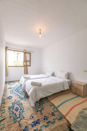 Ein Zimmer mit zwei Betten, einem fensterbelaubten Fenster, einer Hängelampe und einem gemusterten Teppich.