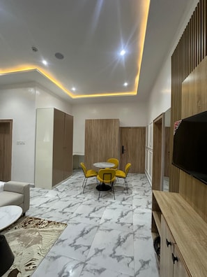 Uma sala de estar moderna com piso de mármore, uma mesa de jantar redonda com cadeiras amarelas, um sofá, uma televisão e painéis de madeira nas paredes.