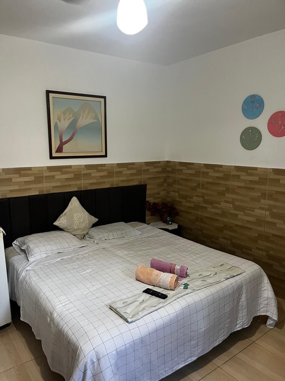 A bedroom with a bed, a nightstand, a wall clock, a framed picture, and a wall-mounted light fixture.