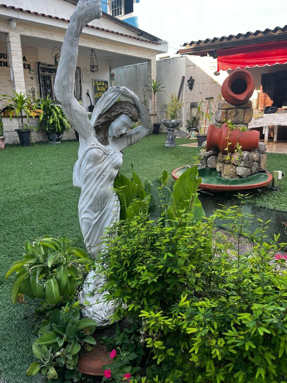 A statue in a garden with a fountain, potted plants, and a building in the background.