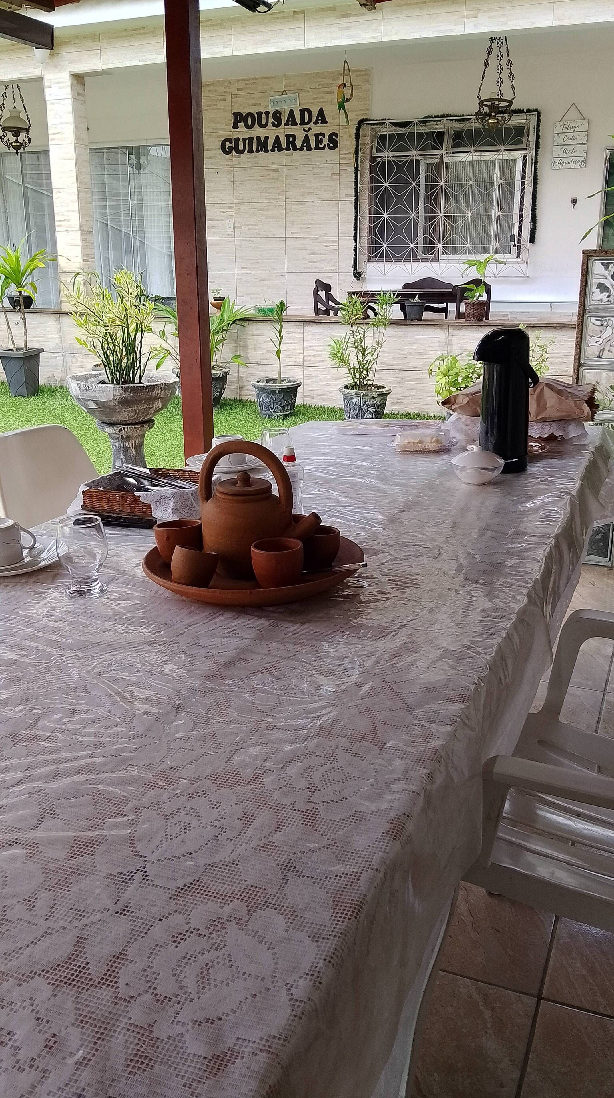 Uma área de refeições coberta ao ar livre, com mesa posta para chá, rodeada por plantas em vasos e um edifício com a placa 'POUSADA GUIMARÃES'.