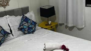 A neatly made bed with a rolled towel and a flower, a bedside lamp, decorative wall art, and a curtain.