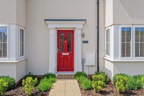 Una casa con puerta roja, detalles blancos y un pequeño jardín con setos podados.