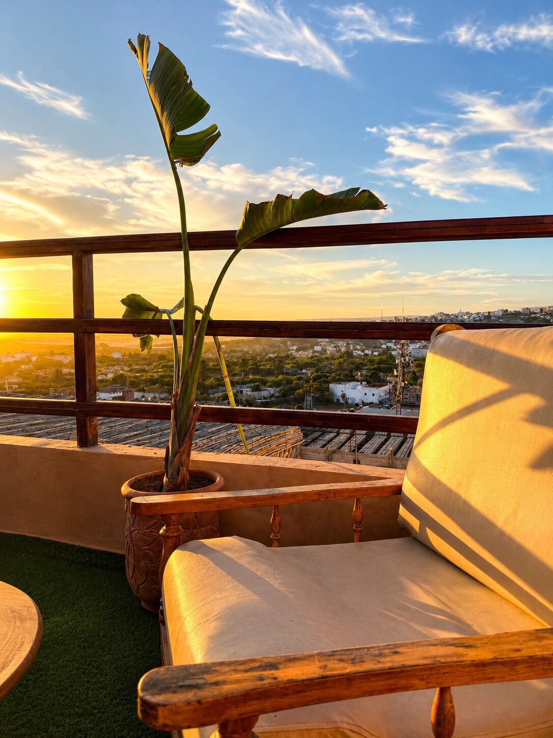 Un balcon agrémenté d’un pot de fleurs, d’un fauteuil confortable et offrant une vue sur le paysage urbain au coucher du soleil.