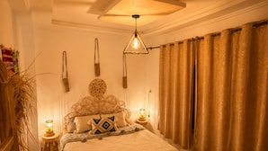 Un dormitorio con cama, mesitas de noche, lámpara colgante, cortinas y una silla.