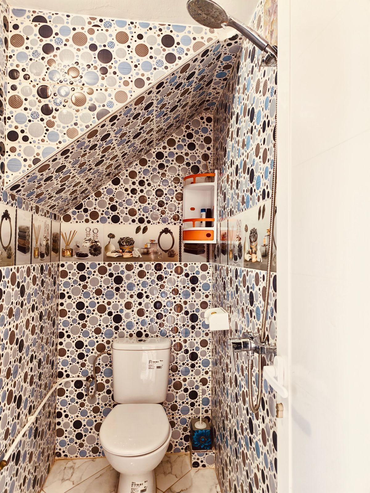 A bathroom with a polka dot tile pattern, a toilet, a shower, and a shelf with toiletries.