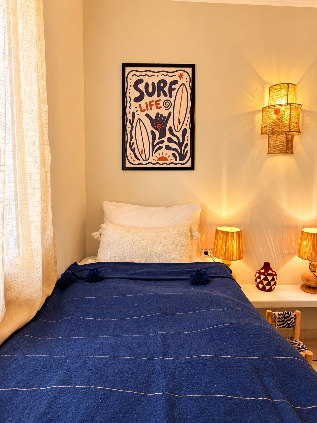 Un dormitorio con una cama, una ilustración enmarcadas de salvavidas, dos lámparas fijadas a la pared y un ventanal con cortinas.