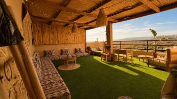A rooftop terrace with a thatched roof, green artificial grass, wooden furniture, and a view of the cityscape.