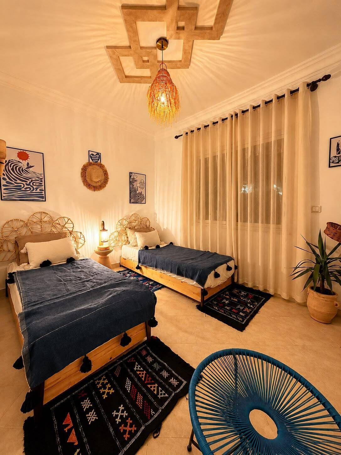 A bedroom with two beds, a hanging light fixture, a potted plant, and a chair.