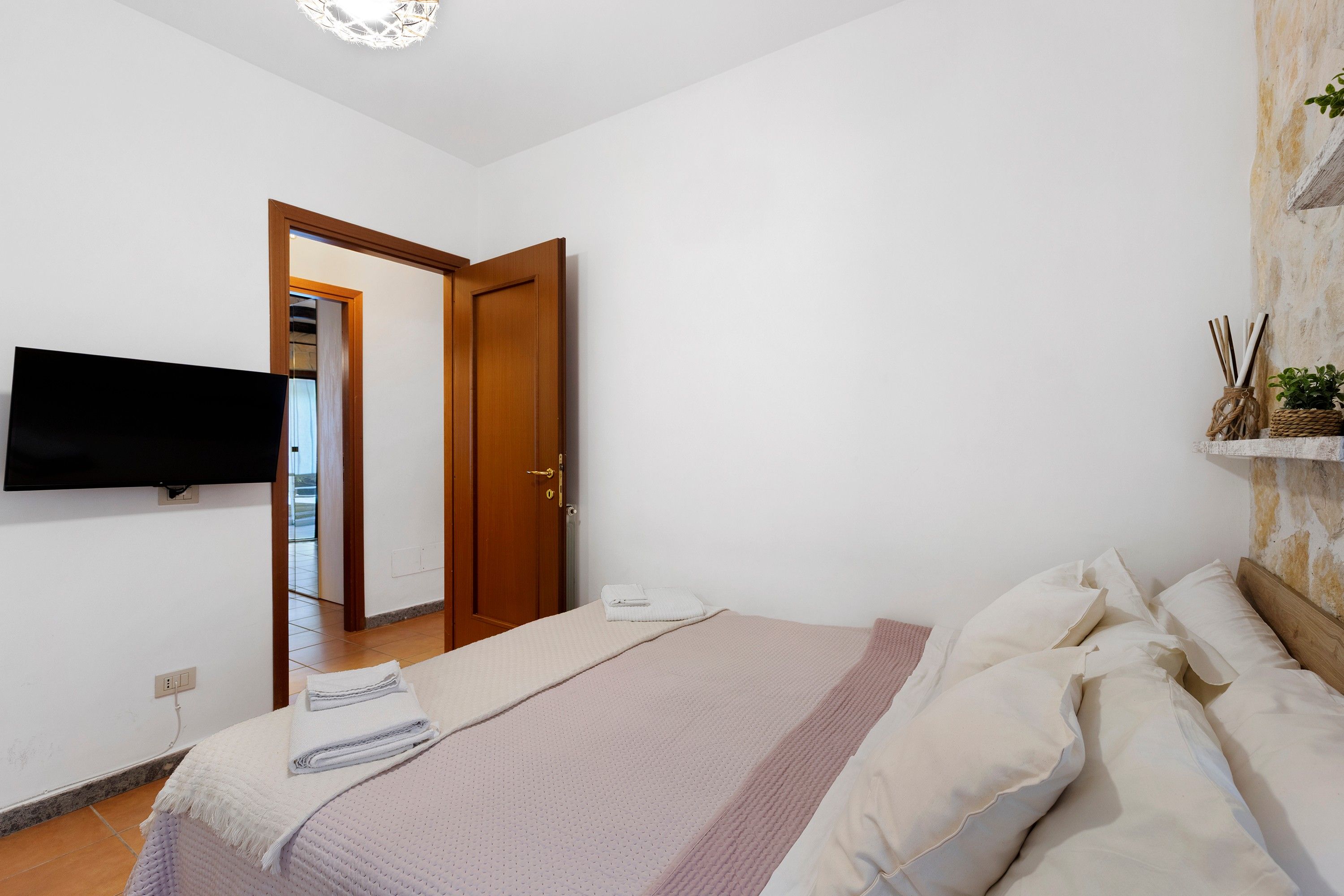 A bedroom with a bed, a flat-screen TV mounted on the wall, a wooden door, and a small plant on a shelf.