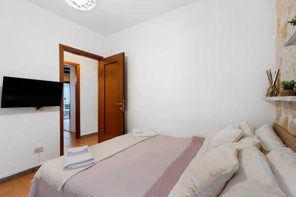 Une chambre à coucher avec un lit, un téléviseur à écran plat fixé au mur, une porte en bois et une petite plante posée sur une étagère.