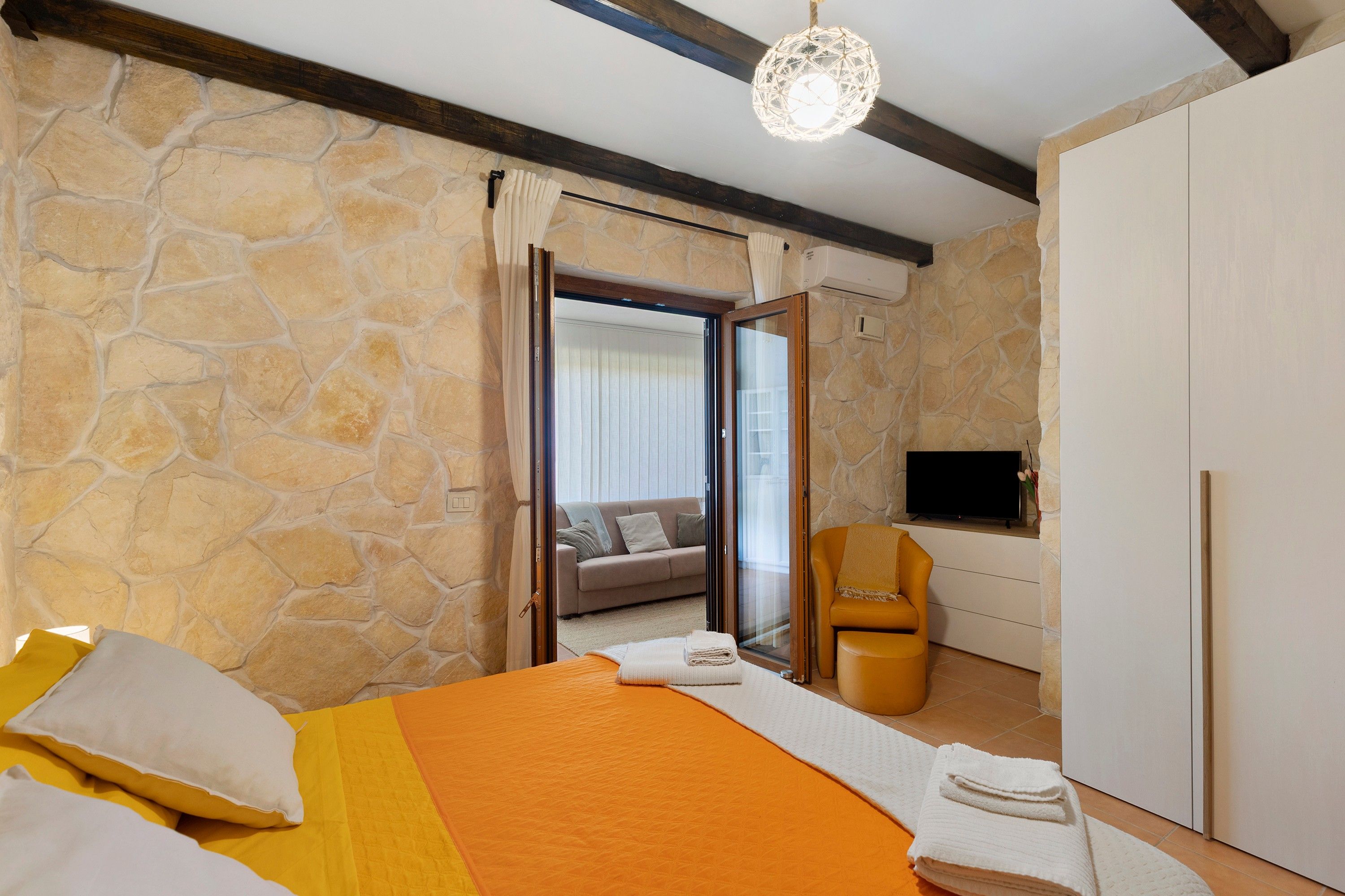 Ein Hotelzimmer mit Steinsandsteinwand, einem Bett mit orangefarbener Bettdecke, einer Couch, einem Fernseher und einer Holzbalkendecke.