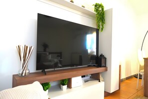 A modern living room with a large flat-screen TV mounted on a wooden console, a white sofa, a small potted plant, and a decorative vase with sticks.