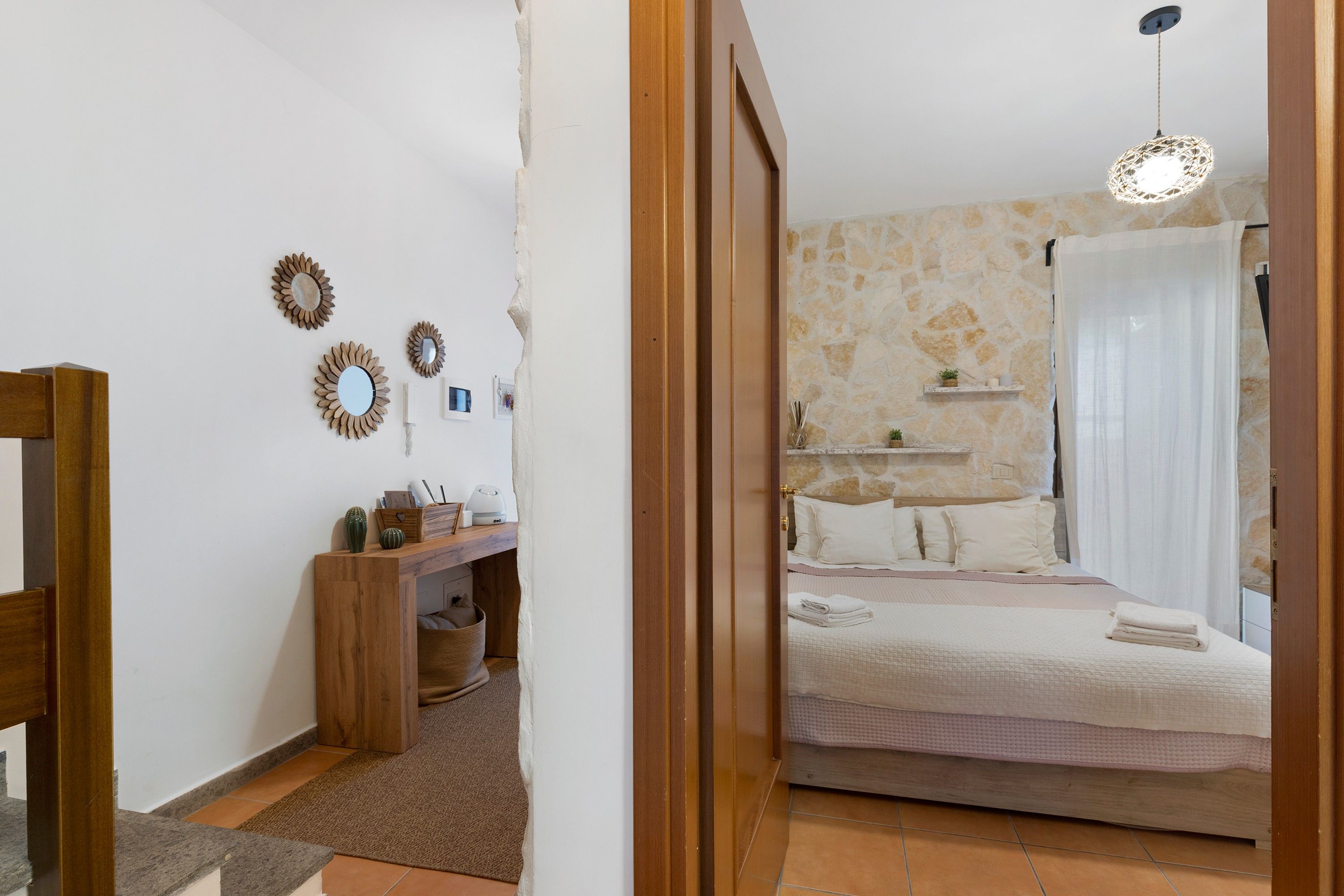 Una camera da letto con parete in pietra, un letto con biancheria bianca, un comodino con una lampada e una scrivania in legno con specchio e un cactus.