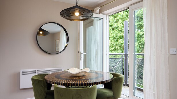 A dining area with a round table, two chairs, a hanging light fixture, a round mirror, and a sliding glass door leading to a balcony. - Oak & Earth Isle of Dogs Apartment (London)