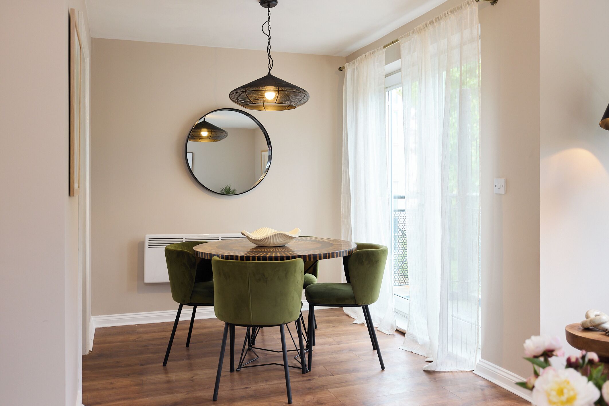 A dining area with a round table, two green chairs, a hanging light fixture, a round mirror, and a window with sheer curtains.
