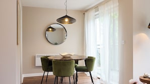 A dining area with a round table, two green chairs, a hanging light fixture, a round mirror, and a window with sheer curtains. - Oak & Earth Isle of Dogs Apartment (London)