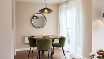 A dining area with a round table, two green chairs, a hanging light fixture, a round mirror, and a window with sheer curtains.