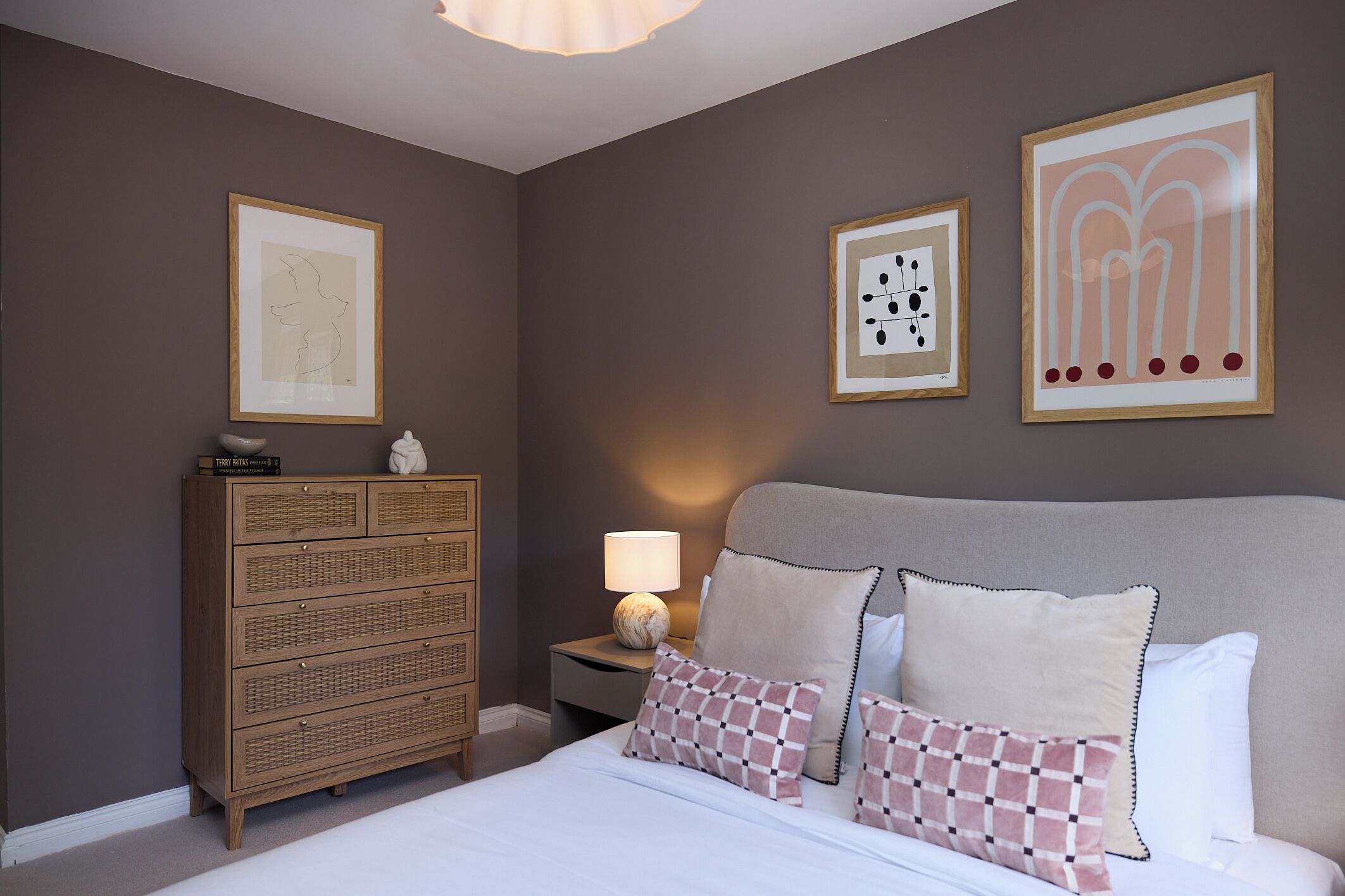 Ein Schlafzimmer mit einem Bett, einer Kommode, einem Nachttisch mit Lampe und drei gerahmten Kunstwerken an der Wand.