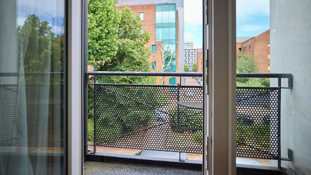 A view through a window with a metal railing, a building with glass windows, and trees in the background. - Oak & Earth Isle of Dogs Apartment (London)
