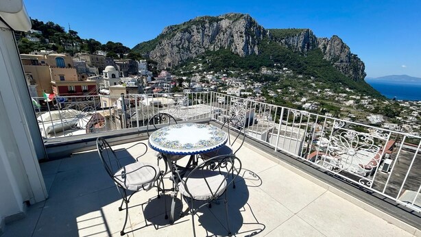 Ein Balkon mit rundem Tisch und Stühlen, mit Blick auf eine Küstenstadt mit Gebäuden und einem Berg.