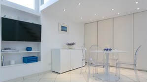 Ein modernes Zimmer mit einem weißen Schrank, einem Flachbildfernseher, einem Esstisch mit klaren Stühlen und einem großen Fenster.