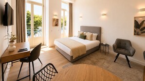 Quarto de hotel moderno com uma cama grande, uma escrivaninha, uma cadeira, uma mesinha e uma TV fixada na parede.