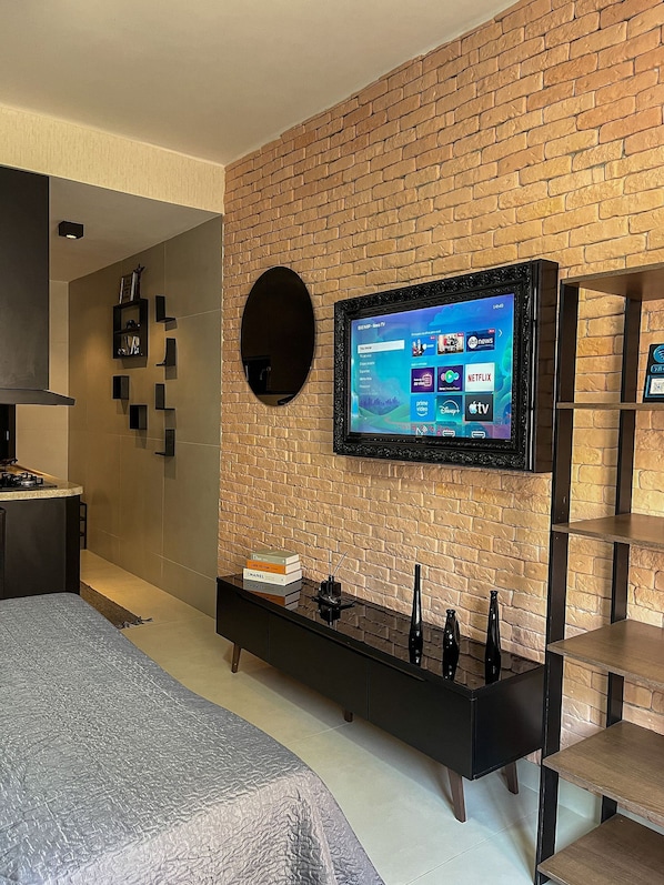 Ein modernes Hotelzimmer mit Backsteinwand, einem flachbildfernseher, der daran befestigt ist, einer schwarzen Unterhaltungseinheit darunter und einem Bett mit grauem Strukturbezug.