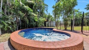 A circular hot tub with a brick border, surrounded by a metal fence, set in a lush garden with tall trees.