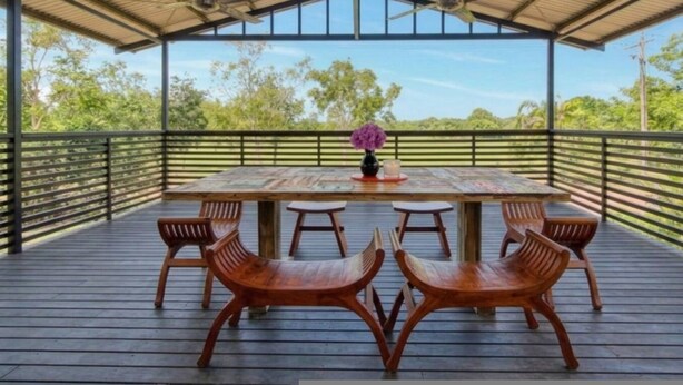 A covered outdoor seating area with a table and chairs, a ceiling fan, and a view of trees.
