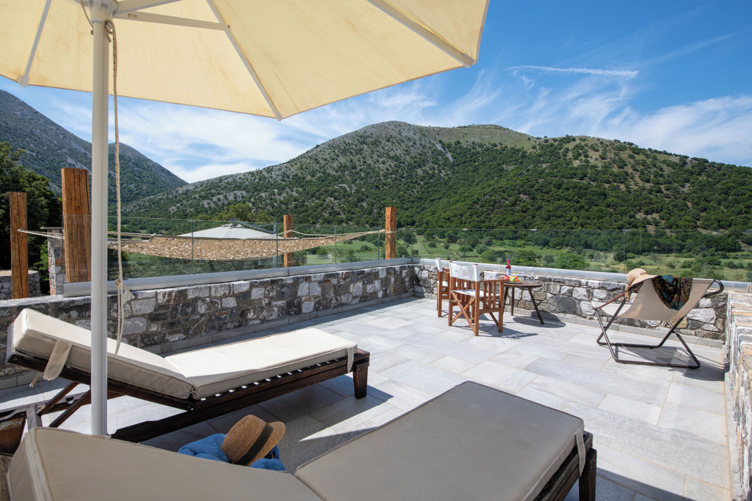 A terrace with a stone wall, a wooden table, a lounge chair, and a large umbrella.
