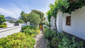 Un étroit sentier mène à un bâtiment blanc doté d’une fenêtre, entouré d’une végétation luxuriante et avec une montagne en arrière-plan.