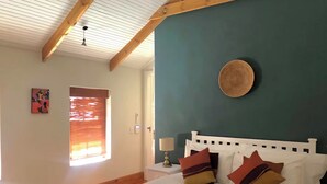 A bedroom with a bed, a nightstand, a lamp, a window with blinds, and a wall-mounted circular object. - Dream Holiday Home in the Heart of Franschhoek (Franschhoek)