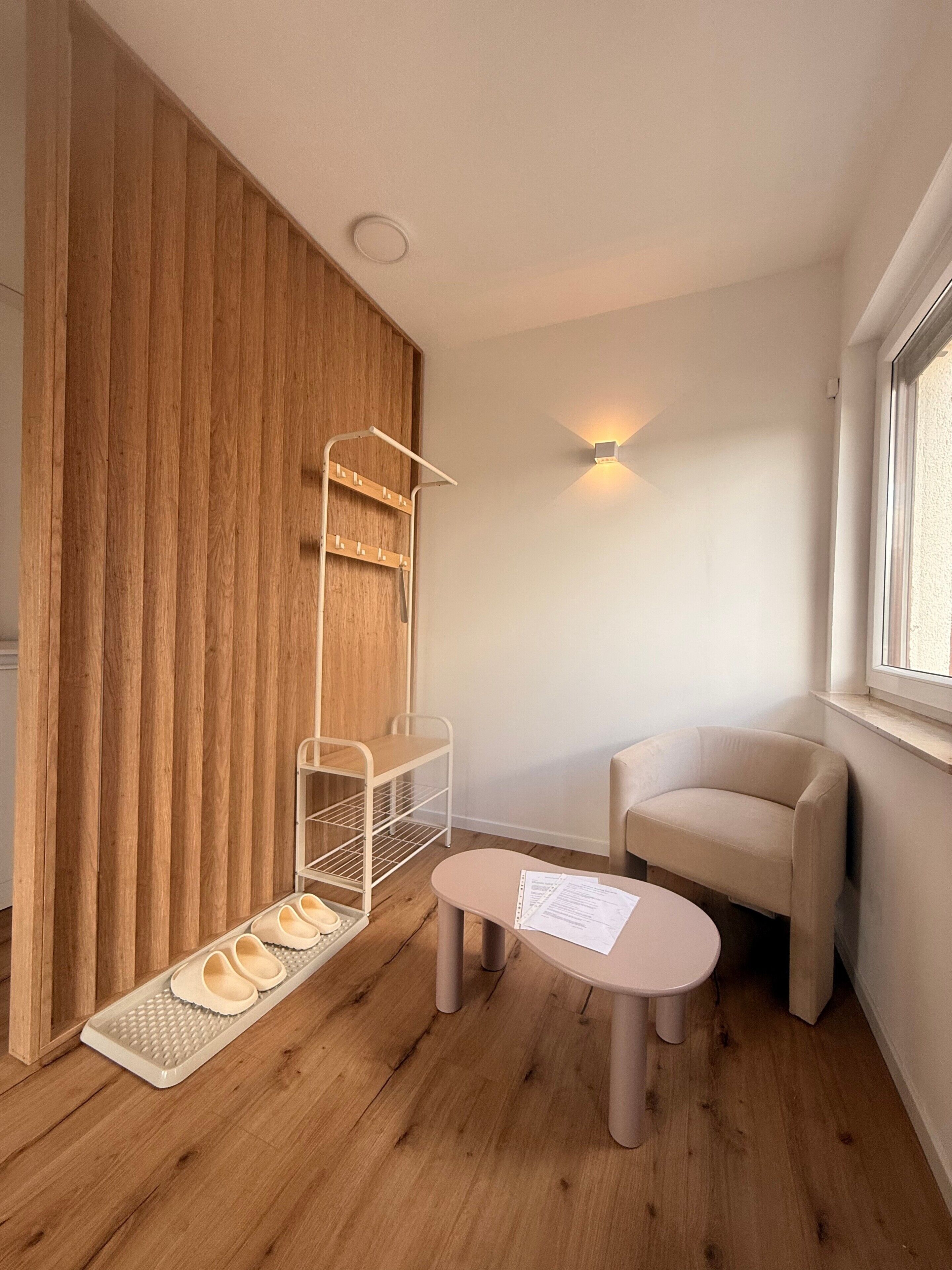 Ein Zimmer mit einer Holzwand, einem weißen Regal, einem beigen Sessel, einem kleinen runden Tisch mit Papieren und einem Fenster.