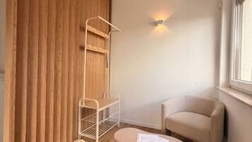 Ein Zimmer mit einer Holzwand, einem weißen Regal, einem beigen Sessel, einem kleinen runden Tisch mit Papieren und einem Fenster.
