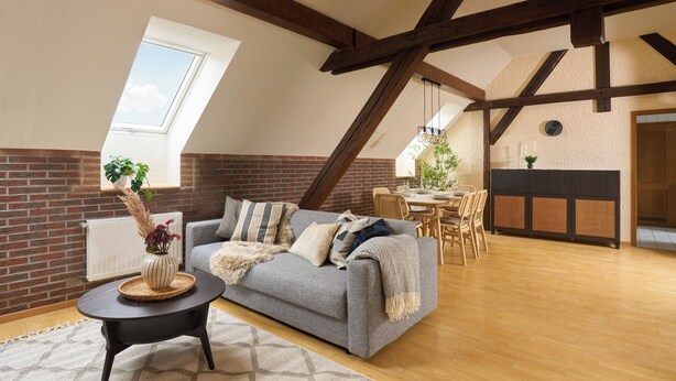 Una habitación estilo loft con vigas de madera a la vista, un sofá gris, una zona de comedor y una pared de ladrillo.