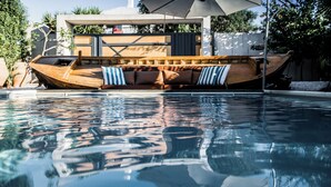 Ein bootsförmiger Pool mit Holzterrasse und Kissen, umgeben von einem modernen Haus und Palmen.