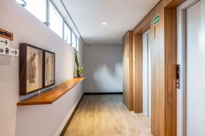A hallway with wooden flooring, framed artwork on the wall, a plant, and a sign indicating the direction to the elevator. - Way Bossa #J5 - Apartamento en Bessa by Carpediem (João Pessoa)