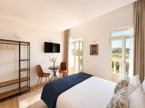 Ein Hotelzimmer mit Bett, Fernseher, Schreibtisch mit Stühlen, Kleiderständer und einem Balkon mit Aussicht.