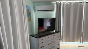 A room with a TV on a wooden cabinet, an air conditioner, white curtains, and a painting on the wall. - Tata Tides Getaway (Tatamagouche)
