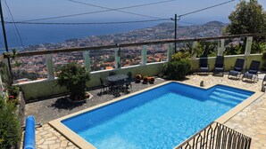 A rectangular pool with a clear blue water, surrounded by stone tiles. There are lounge chairs and a table with chairs. A view of a cityscape with buildings and a body of water is visible through a balcony.